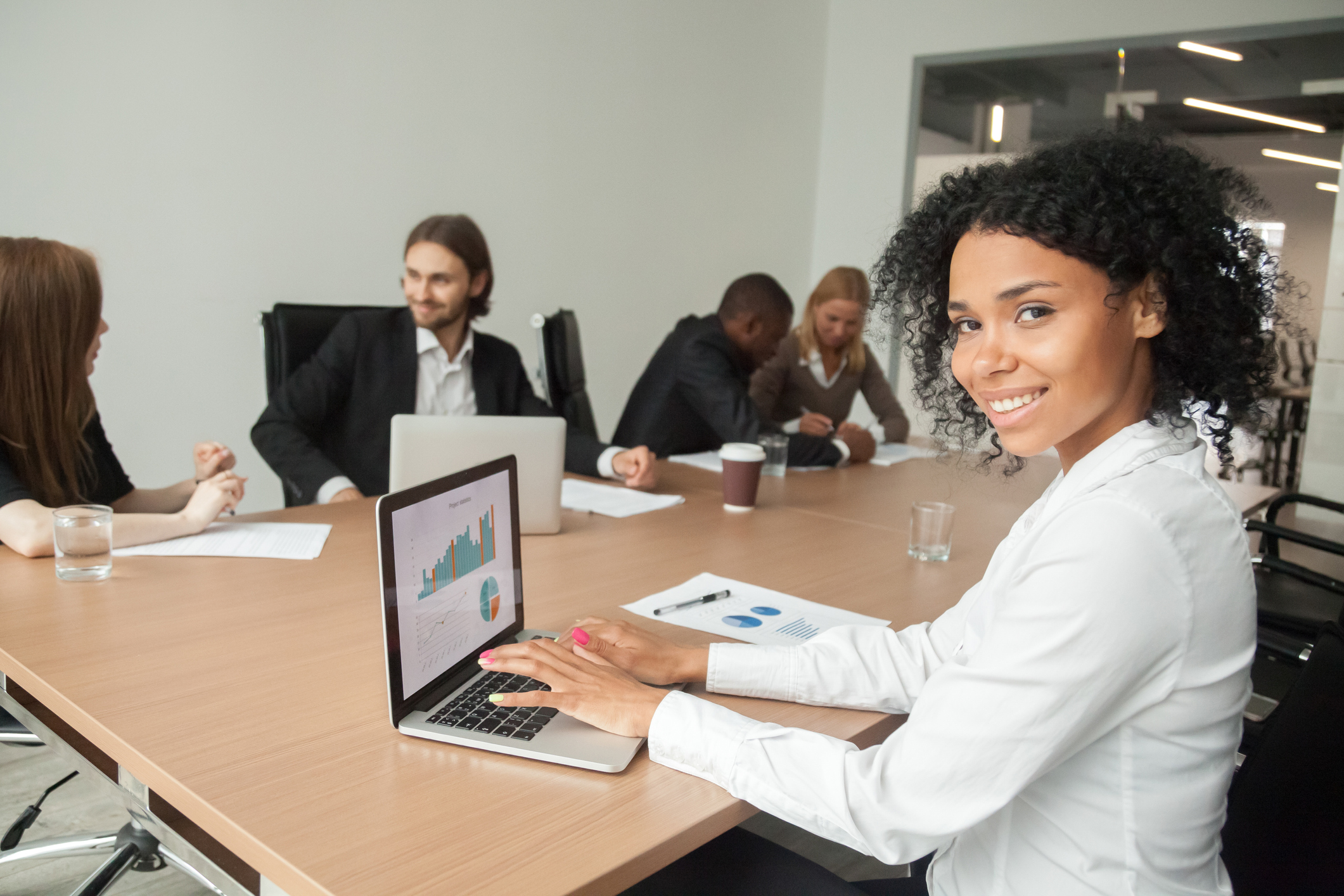 African smiling businesswoman looking at camera using laptop at meeting Smiling millennial african businesswoman looking at camera at meeting, female afro american marketing manager using laptop at group office briefing, young black project sales professional portrait